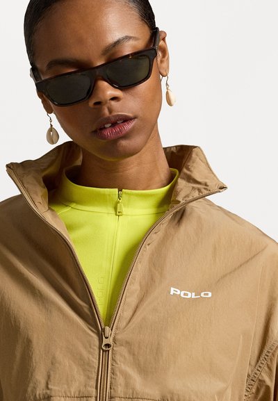 Brūns jaku ar rāvējslēdzēju un paceltu apkakli, apvienots ar spilgti dzeltenu kreklu. Aksesuāri ietver melnus saulesbrilles un čaulu karājas auskari.