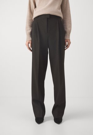 Personne portant un pantalon noir taille haute à jambes larges avec des plis sur le devant et un pull en maille beige, debout devant un fond clair uni.