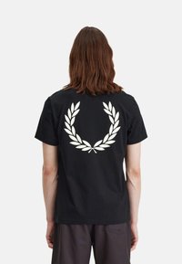 Camiseta corta negra de algodón con un gran gráfico blanco de una corona de laurel en la parte de atrás. Cuello redondo sencillo y corte relajado.