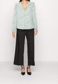 Blouse verte menthe à motif floral avec un décolleté en V, des épaules bouffantes et des manches longues, associée à un pantalon large noir taille haute et des talons noirs.