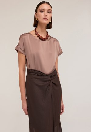 Korte mouw, licht taupe blouse van een zijdezacht materiaal, gecombineerd met een donkerbruine rok met een gedraaide knoopdetail. Ronde houten ketting als accent.