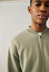 Sudadera de color verde oliva con cremallera, hecha de una tela suave. Presenta un cuello acanalado y una textura lisa, diseñada para ajustarse cerca del cuello.