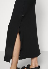 Jupe midi noire avec fente latérale dévoilant le bas de la jambe, portée avec des sandales noires à boucle sur fond clair.