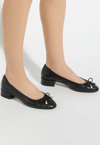 Zwarte leren balletschoenen hebben een ronde neus, een kleine blokhak, een strikdetail met parelaccenten en een geplooide textuur op de bovenkant.