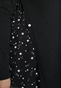 Cardigan noir partiellement ouvert, révélant une chemise noire avec des motifs d'étoiles blanches et de constellations en dessous.