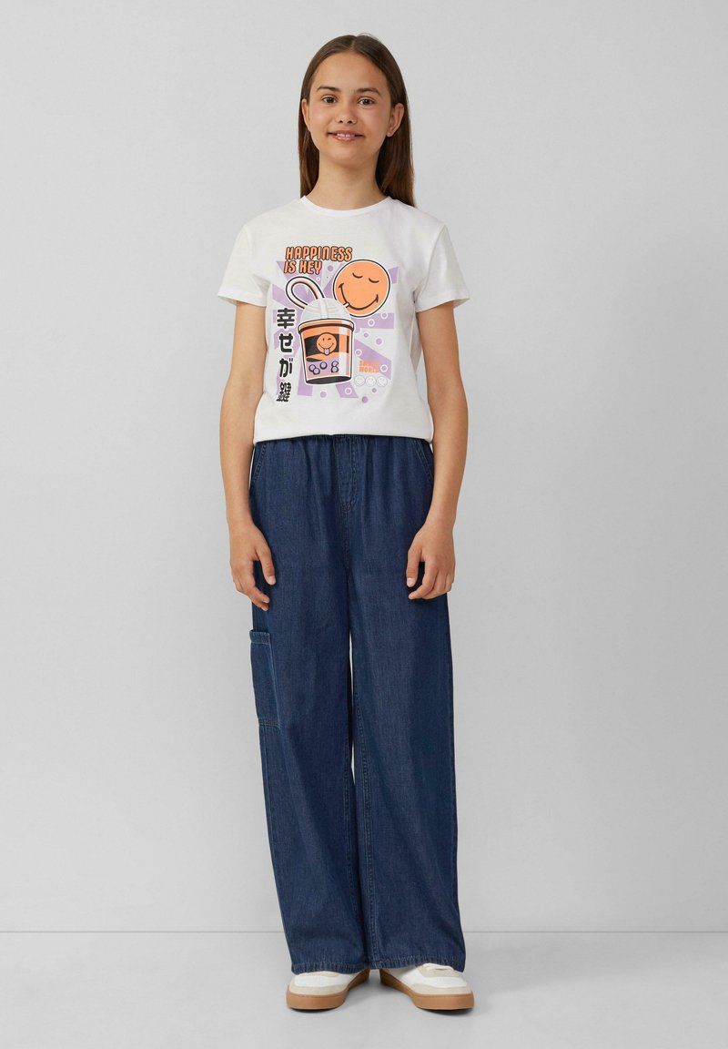 Witte katoen t-shirt met een kleurrijk grafisch ontwerp van een glimlachend gezicht en tekst. Gecombineerd met brede blauwe denimbroek en crèmekleurige sneakers.