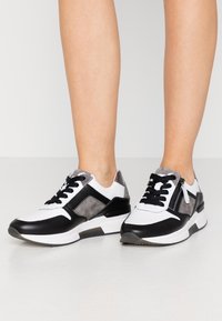 Svarta, vita och grå atletiska skor med en mix av läder och mocka, som har en kontrasterande textur, snörning och en sidodragkedjeförslutning.