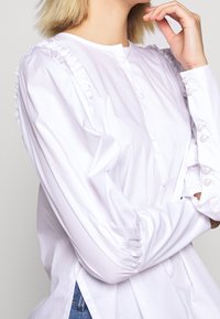 Camicia bianca con bottoni, dotata di dettagli con volant sulle spalle e bottoni in perla sulle maniche. Il tessuto appare liscio e leggermente lucido. Le maniche sono arricciate.