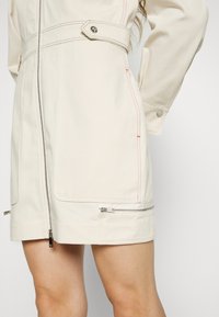 Světle béžová bunda s červeným prošíváním, která má přiléhavý střih, zipové kapsy a vysoký límec se zapínáním na knoflík.