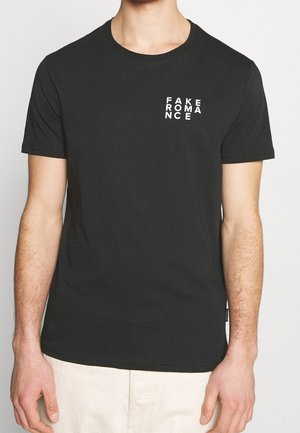 Mann trägt ein schwarzes T-Shirt mit "FAKE ROMANCE" in weißen Blockbuchstaben auf der linken Brust, kombiniert mit hellbeigen Hosen.