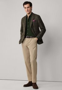 Marynarka w brązową kratę, zielona koszula, beżowe spodnie, brązowe buty. Poszetka dodaje koloru. Gładkie tekstury, dopasowany krój.