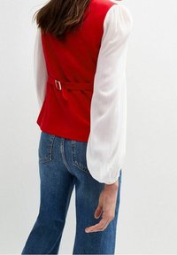 Rote ärmellose Weste mit einem Gürtel in der Taille, kombiniert mit einem weißen, gefältelten Langarmhemd und blauen Jeans. Strukturierter und enganliegender Schnitt.