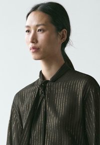 Czarna bluzka ze złotymi pionowymi paskami, z wiązaniem przy szyi i szerokimi rękawami. Gładka tekstura, lekki materiał.