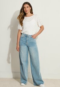 Top de algodón blanco con patrón de ojal y mangas cortas abullonadas, combinado con jeans de tiro alto de pierna ancha en azul claro y zapatillas blancas.