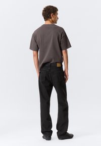 Camiseta de algodón gris oscuro de manga corta, combinada con jeans de mezclilla negros que presentan un parche de cuero y un corte relajado, vista desde atrás.