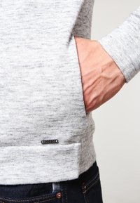 Grauer, strukturierter Pullover mit einer Tasche und geripptem Saum. Verfügt über ein kleines Metall-Logoschild am unteren Ende. Getragen mit dunklen Jeans.