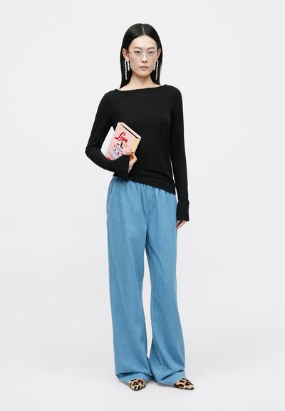 Haut à manches longues noir, jean large bleu clair, lunettes, et chaussures à motif animal. Tenant un livre rose avec une illustration de gâteau.