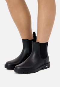 Botas de tobillo de goma negra con paneles laterales elásticos y suelas texturizadas. Acabado liso con una pestaña para facilitar su colocación.