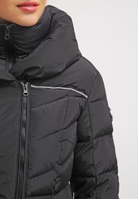 Chaqueta de plumas negra con un cuello alto, que presenta paneles cosidos, un cierre frontal de cremallera y una raya de acento plateada en el bolsillo del pecho.