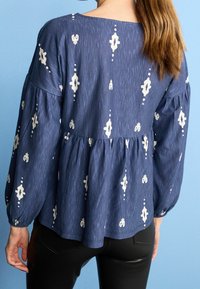 Kobieta ubrana w granatową bluzkę z długim rękawem w białe geometryczne wzory oraz czarne dopasowane spodnie, stojąca na tle jasnoniebieskim.