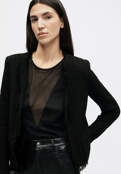 Žena s dlhými tmavými vlasmi, oblečená v čiernom textúrovanom saku, priehľadnom čiernom topu a vysoko pásových čiernych kožených nohaviciach, pózujúca s jednou rukou na bokoch.