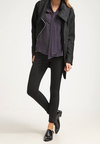 Donna che indossa pantaloni neri su misura, giacca nera a trama, camicetta blu navy a motivi e scarpe eleganti nere lucide, in piedi con le gambe incrociate.