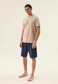 Jasno beżowy bawełniany t-shirt z czarnym napisem i krótkimi rękawami, w zestawieniu z ciemnoniebieskimi spodenki w wzory. Model stoi boso, zwrócony twarzą do przodu.