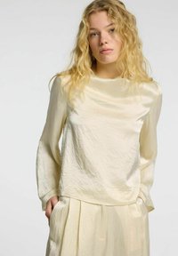 Hellbeige, leichtes Blusenshirt mit einer glatten, leicht gekrümmten Textur und langen Ärmeln. Mit einem runden Ausschnitt und lockerer Passform.