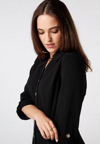 Zwarte jurk met een op maat gemaakt ontwerp, voorzien van gouden knoopaccenten en opgerolde mouwen. Gladde textuur met een gestructureerde pasvorm.