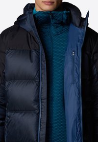 Doudoune bleu marine avec une capuche noire et des côtés noirs, présentant une texture lisse. En dessous, une veste zippée bleu sarcelle avec un motif à pois et des fermetures éclair accentuées.