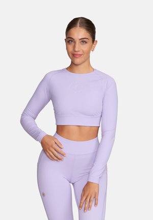 Młoda kobieta związała włosy do tyłu, ubrana w dopasowany lawendowy krótki top z długim rękawem i legginsy z wysokim stanem, stoi z jedną ręką na biodrze.