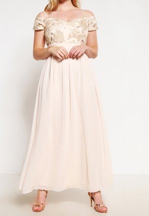 Femme en robe rose clair décolletée aux épaules avec corsage brodé de fleurs, jupe plissée et sandales à talons hauts dorées, debout les mains jointes.