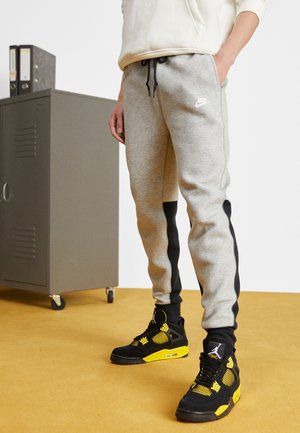 Osoba nosząca szare spodnie dresowe Nike z czarnymi bocznymi panelami oraz czarno-żółte buty Air Jordan, stojąca na musztardowej podłodze obok szarej szafki.