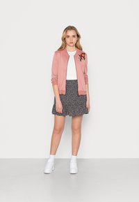 Casaco bomber rosa claro com bordado floral, t-shirt branca e saia floral azul-marinho; modelo vestindo ténis brancos e meias.