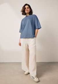 Modré bavlnené oversized tričko v kombinácii s béžovými širokými ľanovými nohavicami a bielymi teniskami s čiernymi doplnkami na neutrálom pozadí.