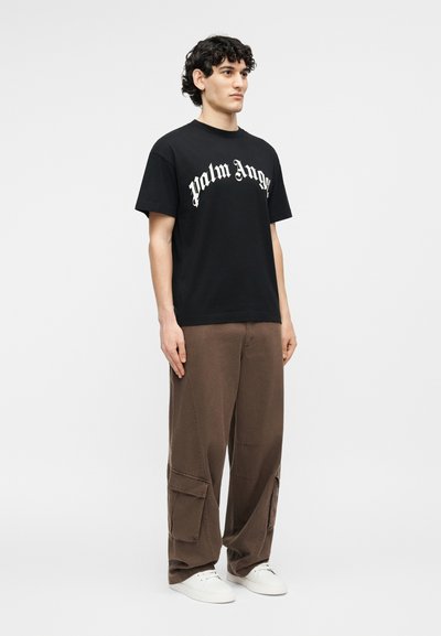 Άνδρας που φοράει μαύρο t-shirt με το λογότυπο "Palm Angels", καφέ παντελόνι cargo και λευκά αθλητικά παπούτσια, στέκεται μπροστά σε ένα απλό ανοιχτό φόντο.