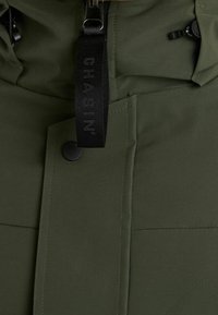 Giacca imbottita verde oliva con collo alto, chiusura con zip, dettagli neri e una vistosa fascia con marchio sulla parte anteriore.