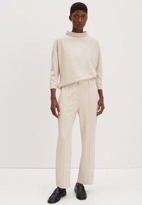 Beige pullover met een hoge kraag, gecombineerd met beige broeken met een rechte pijp en subtiele zijnaad, aangevuld met zwarte instapschoenen.