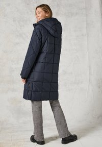 Abrigo acolchado azul marino con capucha desmontable, diseño de rombos, dos cremalleras laterales y detalles en negro. Llevado sobre pantalones gris claro.