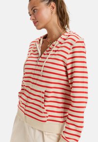 Sudadera de rayas en color crema y rojo, con cierre de cremalleras en la parte delantera, capucha con cordón, bolsillos frontales y dobladillo acanalado. Tejido suave con un corte relajado.
