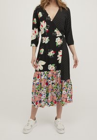 Černé květinové zavinovací šaty s puntíkovými akcenty, tříčtvrtečními rukávy a sukní s barevným květinovým potiskem v několika vrstvách. Stylizováno s bílými teniskami.