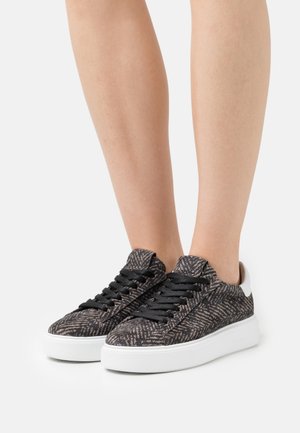 Paire de jambes portant des baskets noires et grises à motifs avec des semelles blanches épaisses et des lacets noirs, debout sur un fond blanc uni.