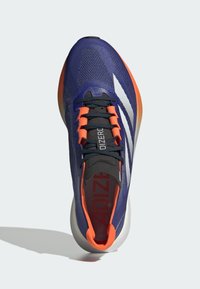 Lilla atletisk sko med orange detaljer, mesh-overdel, sorte snørebånd og tre hvide striber. Har en polstret tunge og en dæmpende sål.
