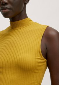 Mosterdgele mouwloze top met een hoge mock kraag, featuring verticale ribbelstructuur en een aangesloten silhouet.