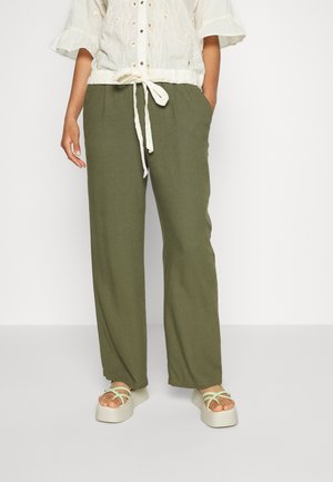 Persoon met olijfgroene wijde broek met trekkoord in de taille, crèmekleurige geborduurde blouse en lichtgroene plateausandalen.