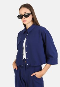 Mörkblå kort kappa med krage, tryckknappar och sidofickor, kombinerad med en vit topp och matchande mörkblå byxor.