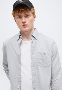 Jeune homme aux cheveux roux bouclés portant une casquette noire, une chemise gris clair à boutons sur un t-shirt blanc, sur un fond clair uni.