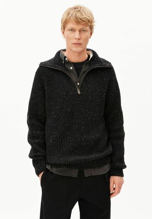 Junger Mann mit blondem Haar trägt einen schwarzen gesprenkelten Zip-Up-Pullover über einem dunklen Hemd mit Knöpfen und schwarzen Hosen vor weißem Hintergrund.