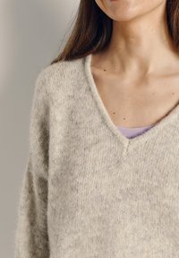 Gaiši bēšs V veida kakla džemperis no mīksta, pūkaina materiāla ar raibu tekstūru, valkāts pār lavandas krāsas topu, ar minimālu plecu laukuma redzamību.