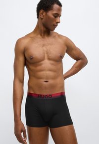 Shorts boxer noirs avec une ceinture dégradée bordeaux arborant le mot "HUGO" en noir. Le tissu semble lisse et ajusté.